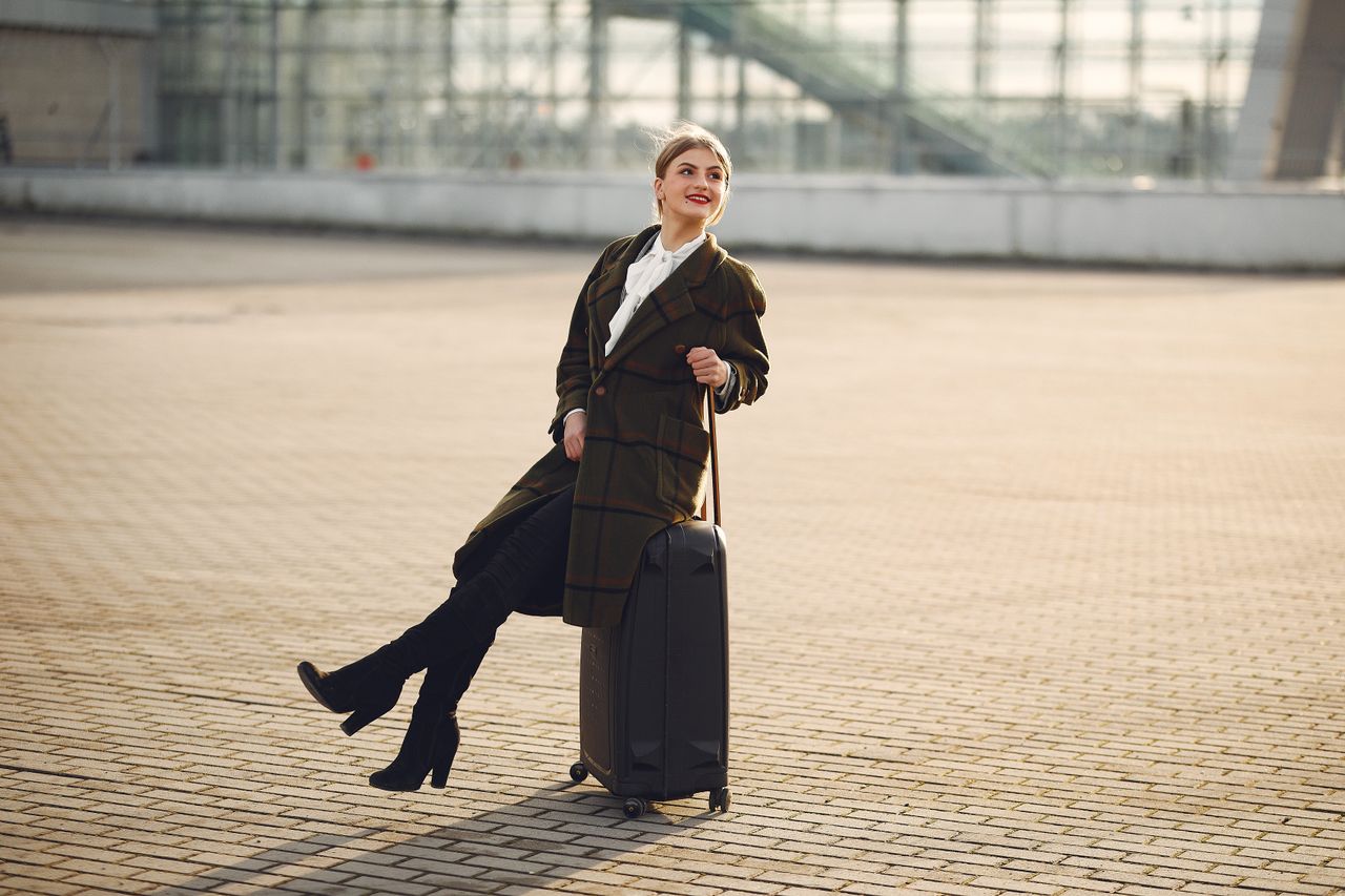 Services Full length of happy female traveler wearing warm trendy plaid coat and high boots sitting on top of suitcase on pavement near modern airport terminal and smiling looking away during daytime in cold season
