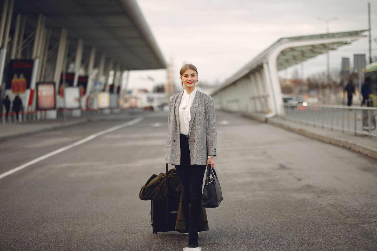 Services Full body of young woman in stylish outfit carrying suitcase and bag while walking along pavement near modern airport building