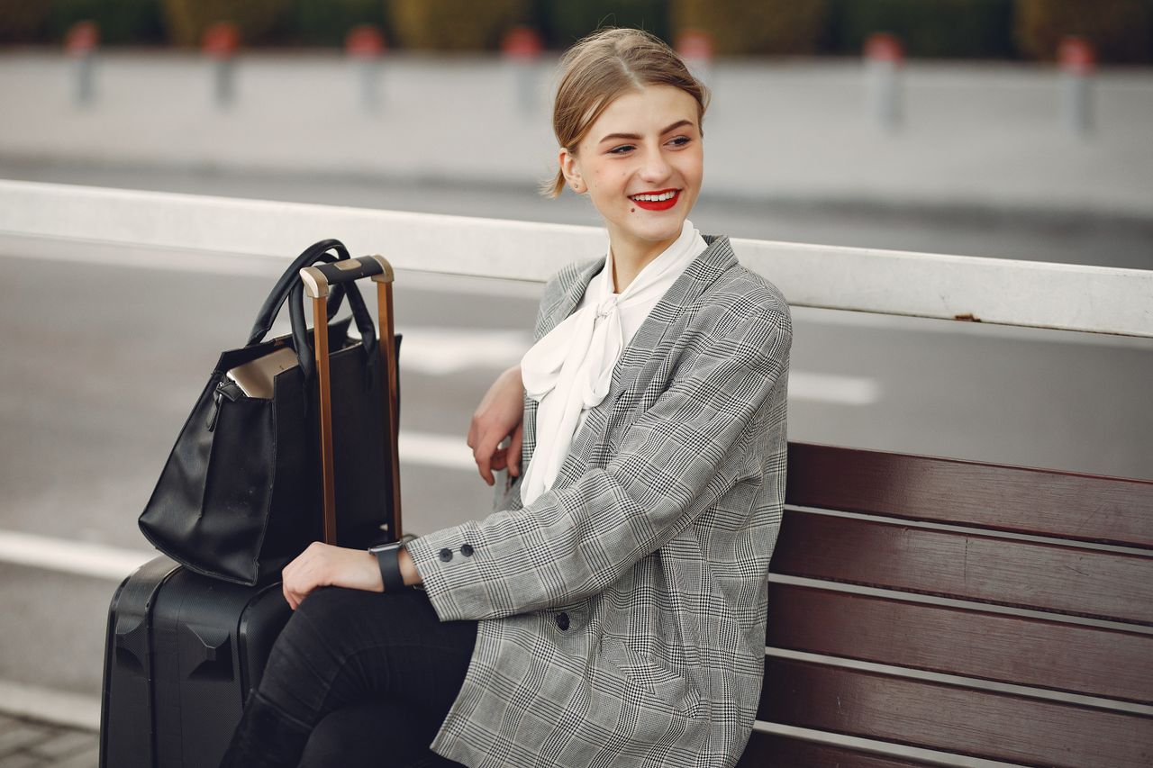 Services Positive female traveler in trendy clothes smiling away while waiting for train sitting on wooden bench with suitcase and bag in street