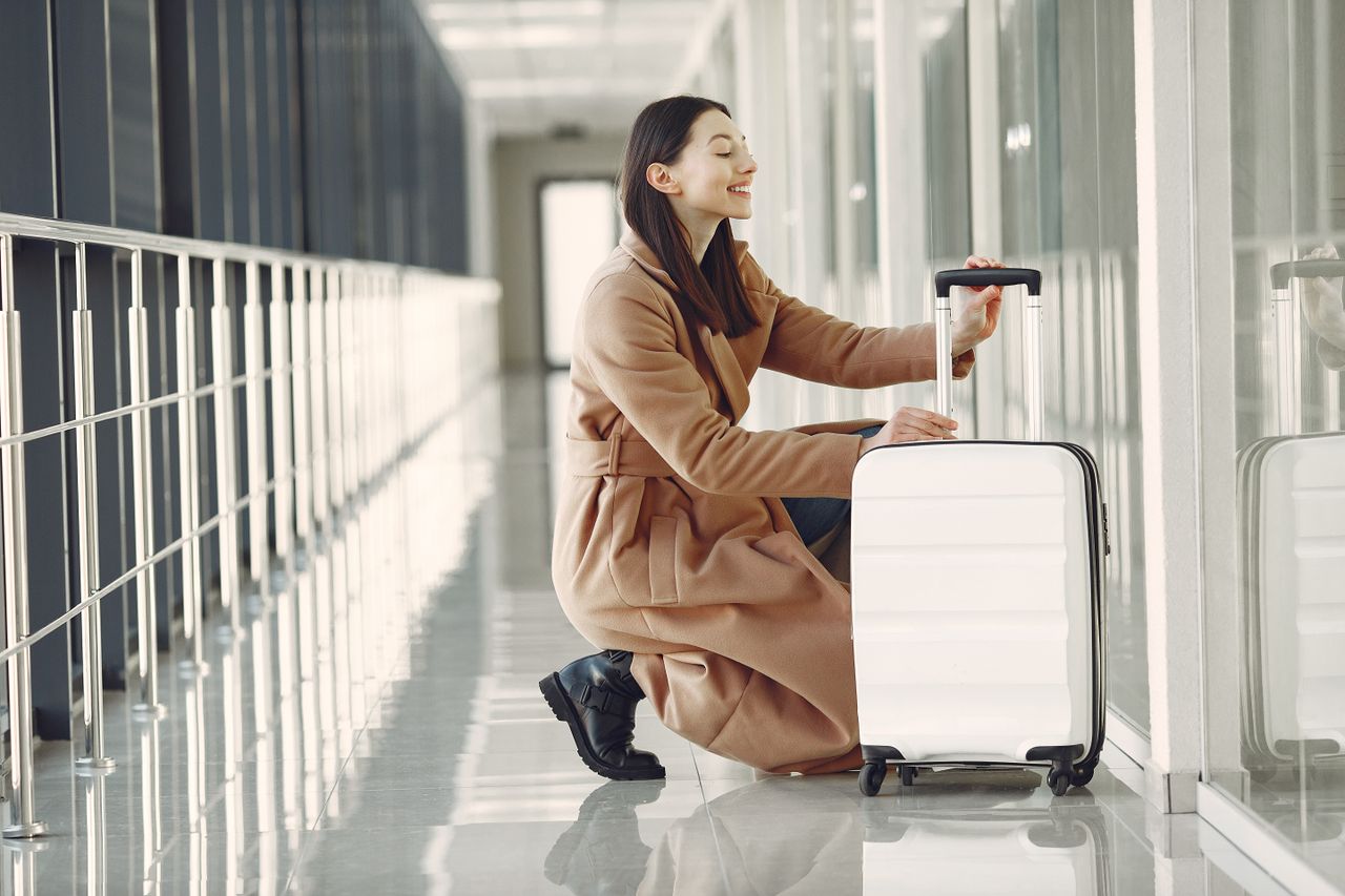 Crafting Captivating Headlines: Your awesome post title goes here Side view full length of cheerful young female passenger in stylish beige coat with suitcase hunkering down and looking out glass wall in contemporary airport corridor