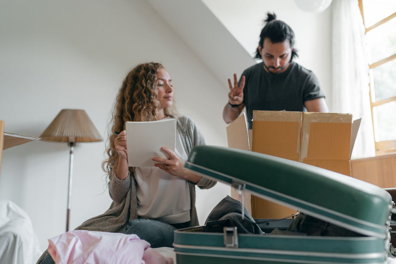 Home Positive concentrated couple counting belonging and taking notes while packing stuff into carton boxes and suitcases near window in sunny bedroom