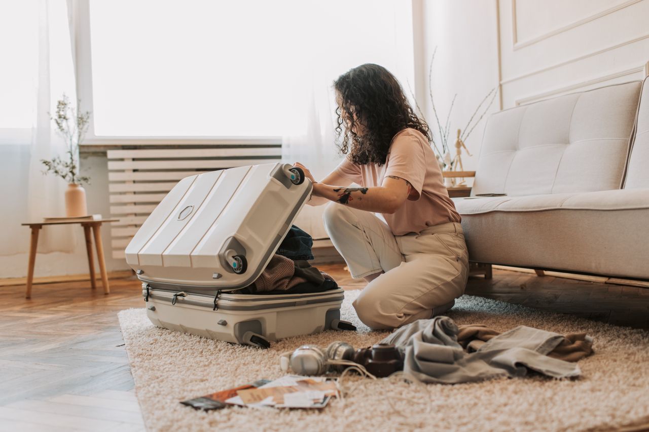 Home A woman packing her suitcase in a stylish living room, surrounded by clothes and travel accessories.